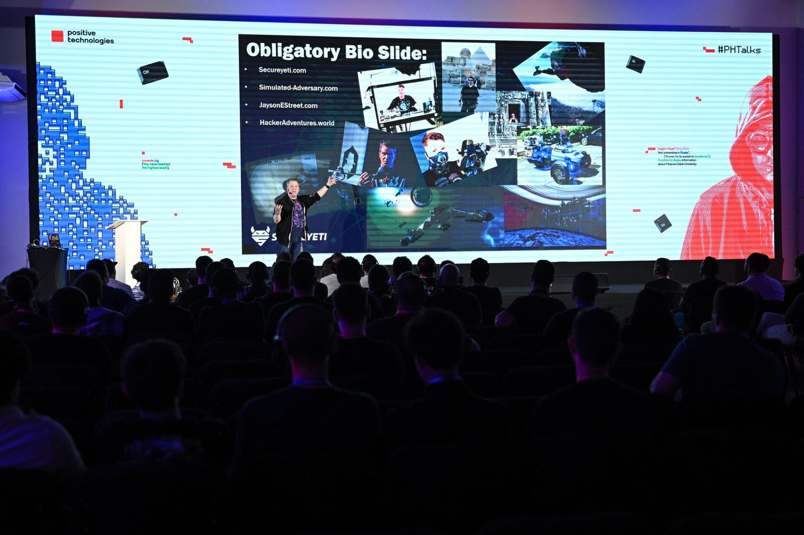 A speaker's bio slide on the big screen in front of a full hall