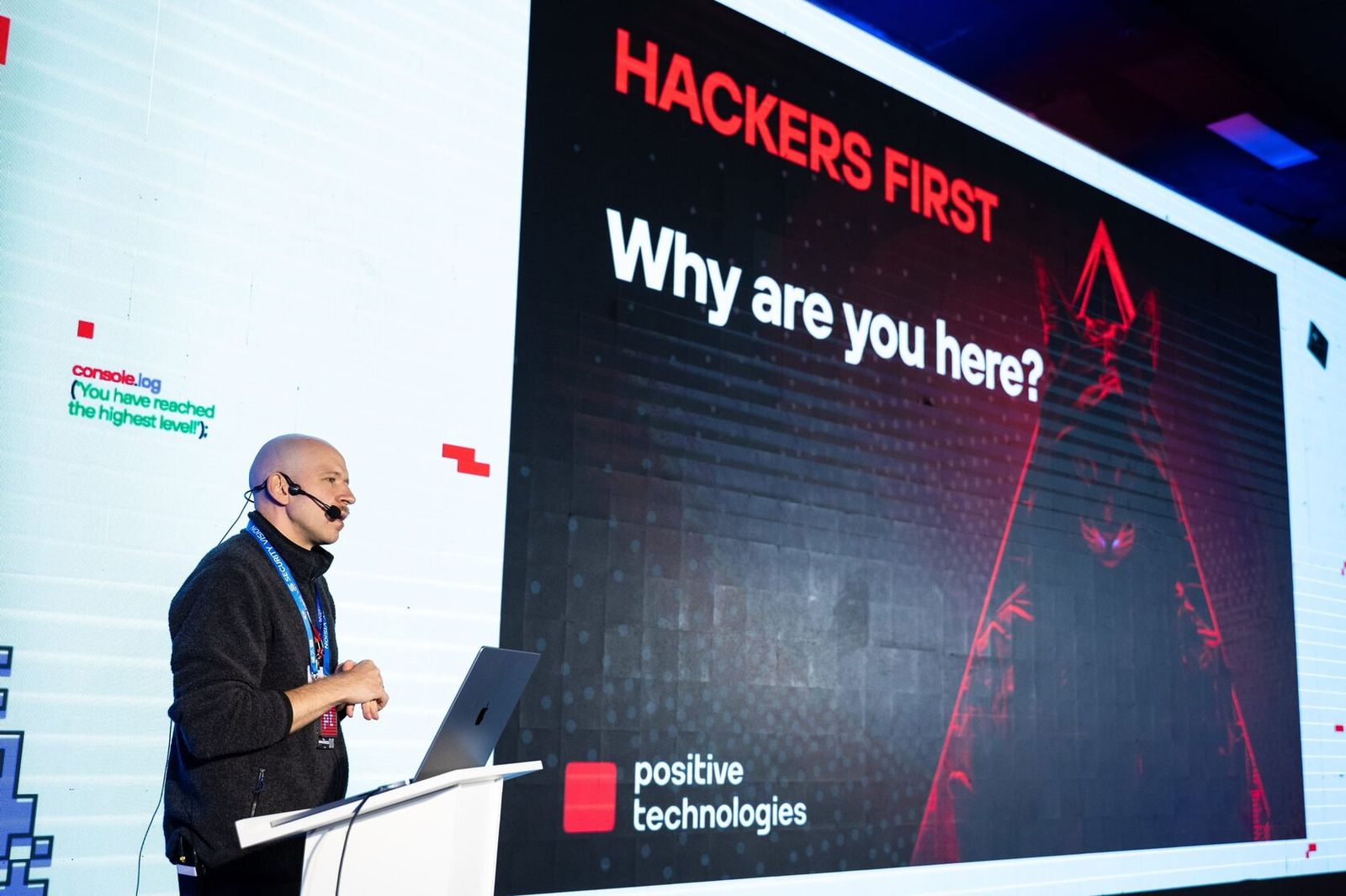 A speaker at the lectern, with "Hackers first. Why are you here?" on the screen